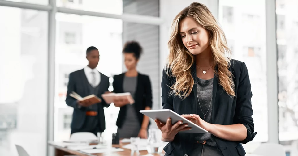 Una donna consulta un tablet con un’espressione concentrata, mentre due colleghi discutono sullo sfondo in una sala riunioni luminosa.
