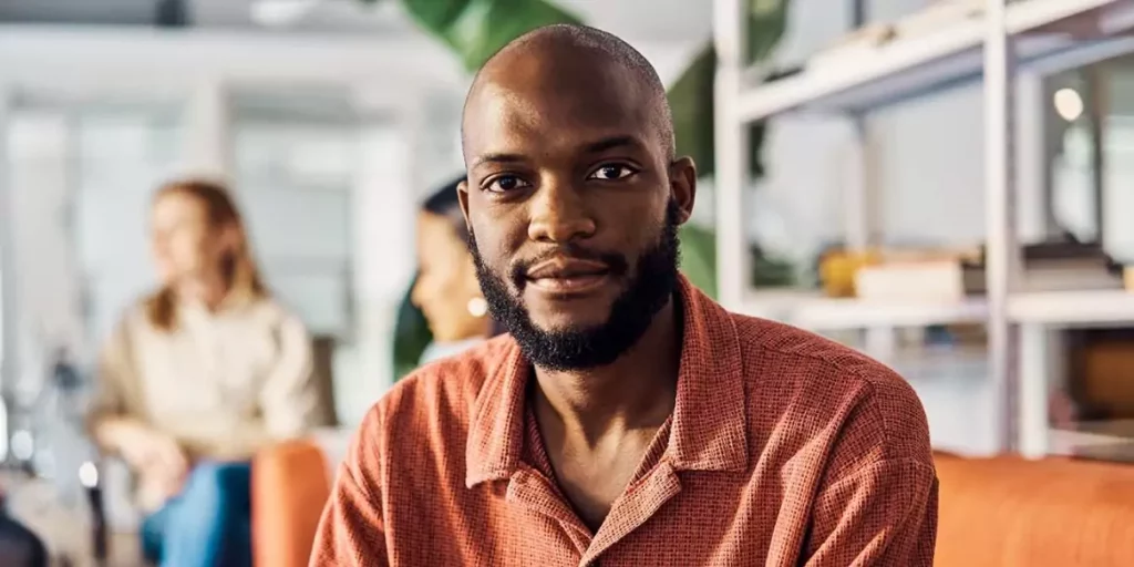 Un uomo guarda verso la fotocamera con un’espressione calma e sicura, seduto in un ambiente di lavoro moderno e informale. 